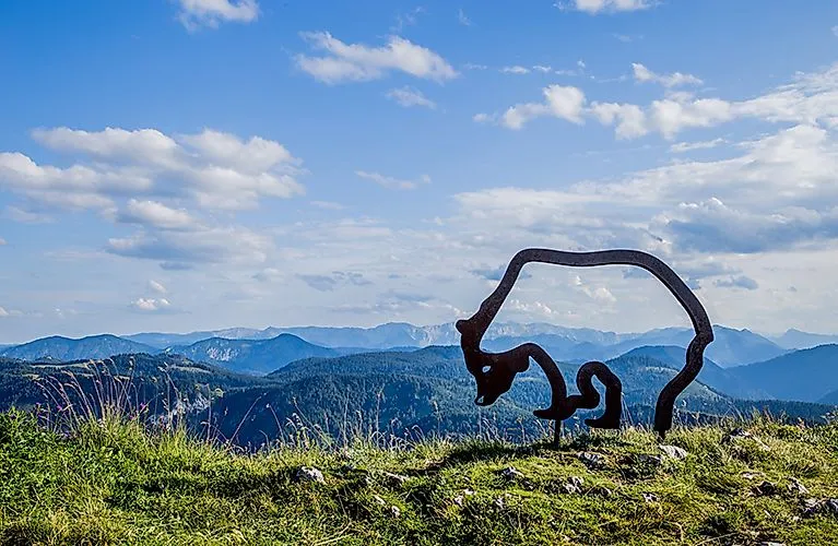 Zomerpanorama bij Lackenhof am Ötscher met modern beeldhouwwerk en uitzicht op beboste heuvels en bergen onder een blauwe hemel.
