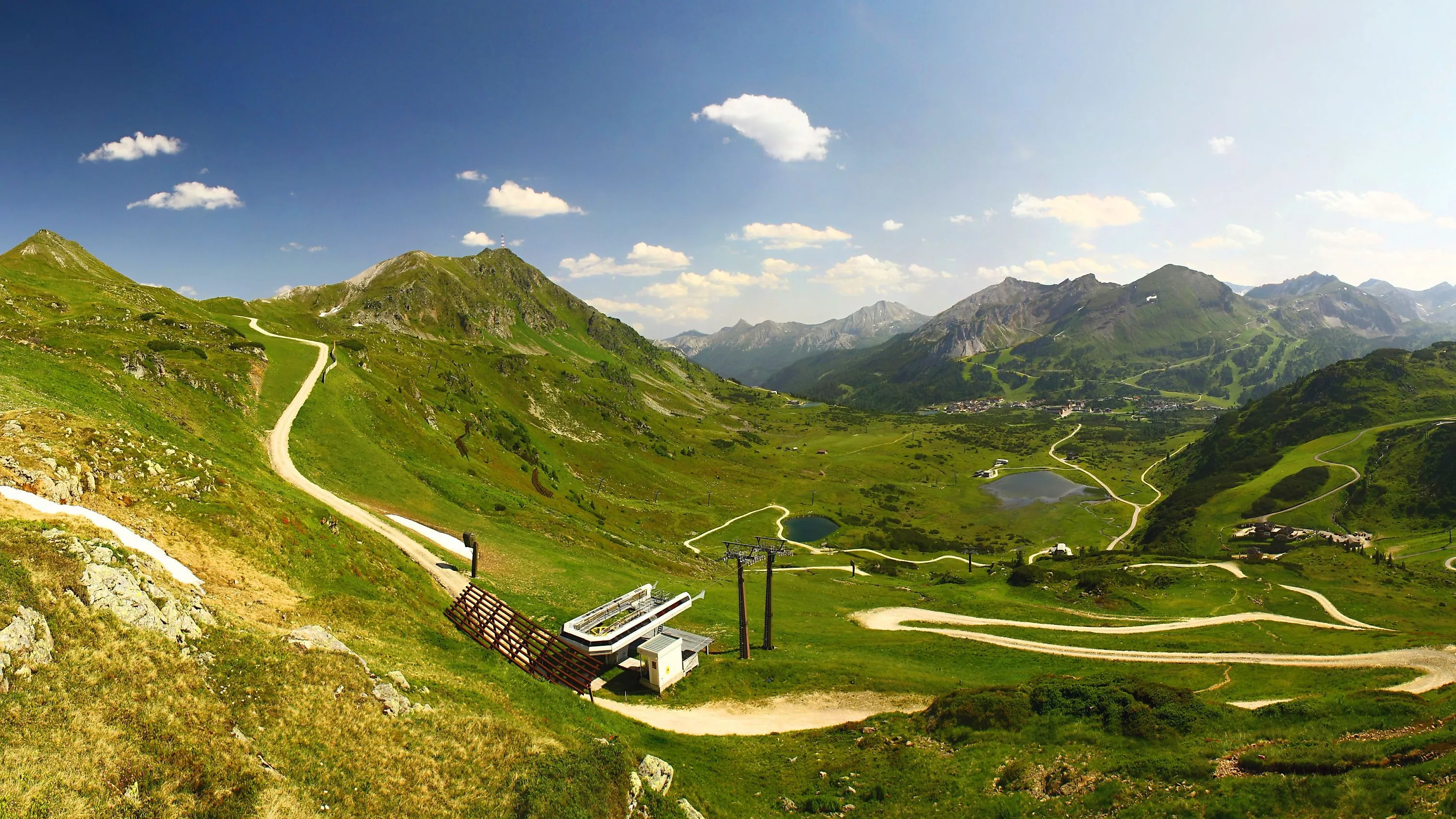 Letní panoramatický záběr na Obertauern s mírnými alpskými loukami, klikatými turistickými stezkami a průzračným horským jezerem, zasazeným do impozantního horského světa Radstädter Tauern.