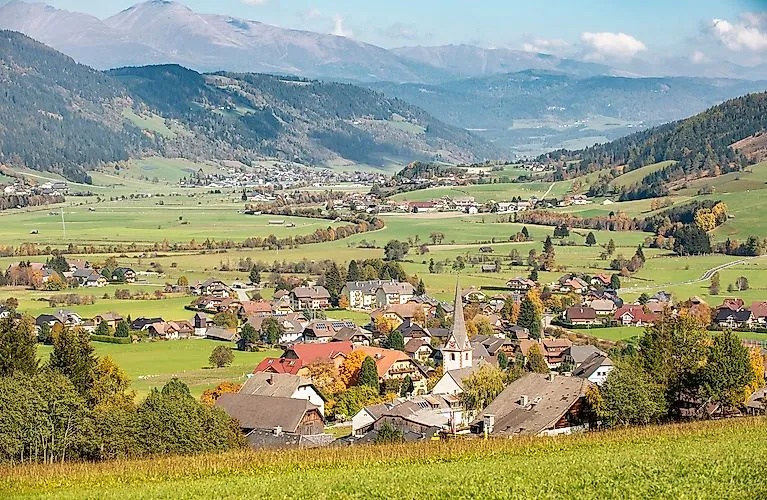 Daleký výhled na zelenou krajinu St. Margarethen im Lungau s roztroušenými domy, poli a horami v pozadí.