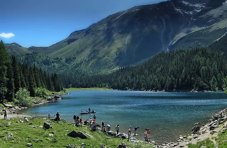 Lidé si užívají letní den u idylického horského jezera nedaleko Steinachu v Tyrolsku, obklopeni zelenou horskou krajinou.